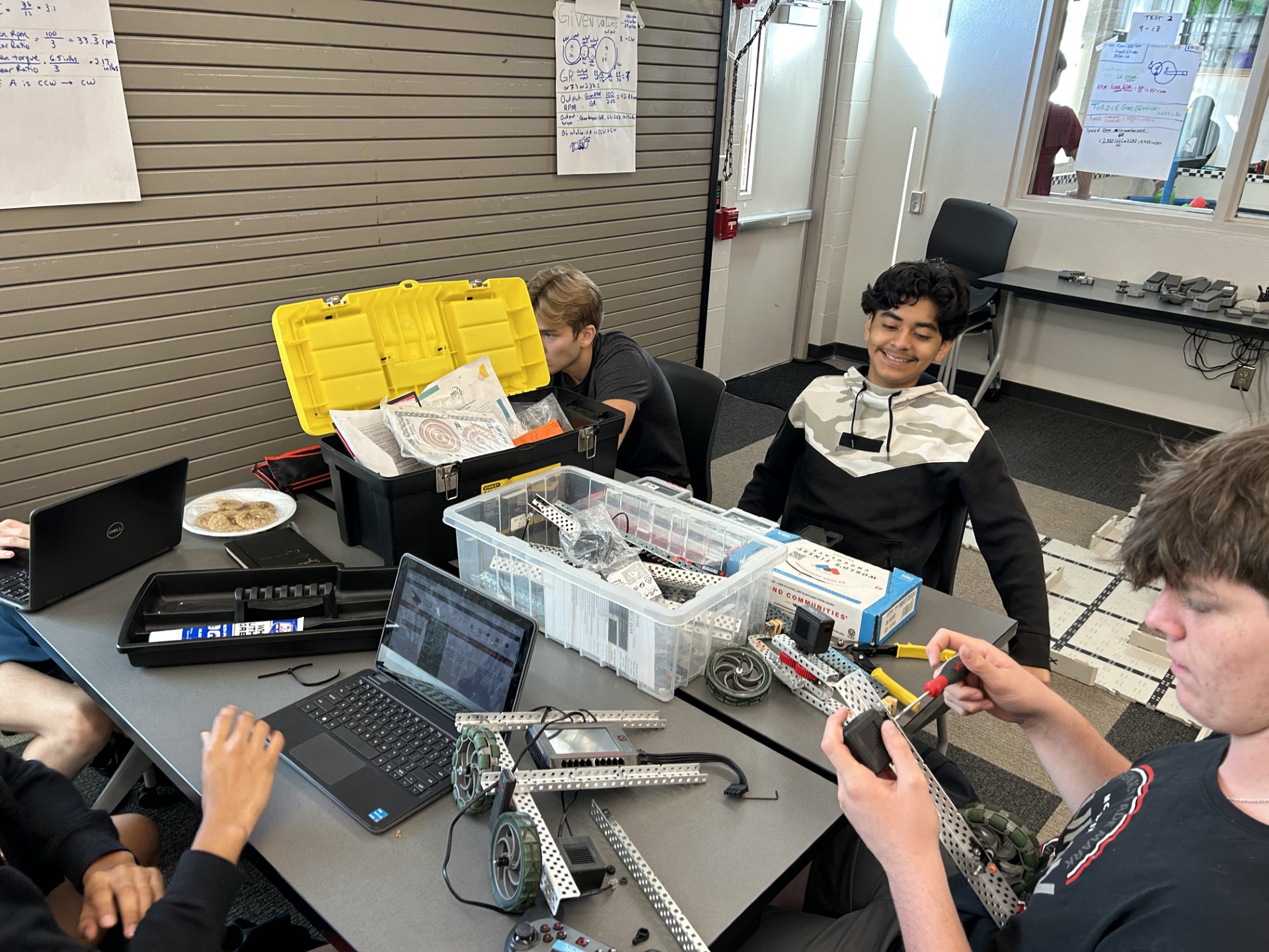 A group of people sitting at a table with laptops and electronicsDescription automatically generated